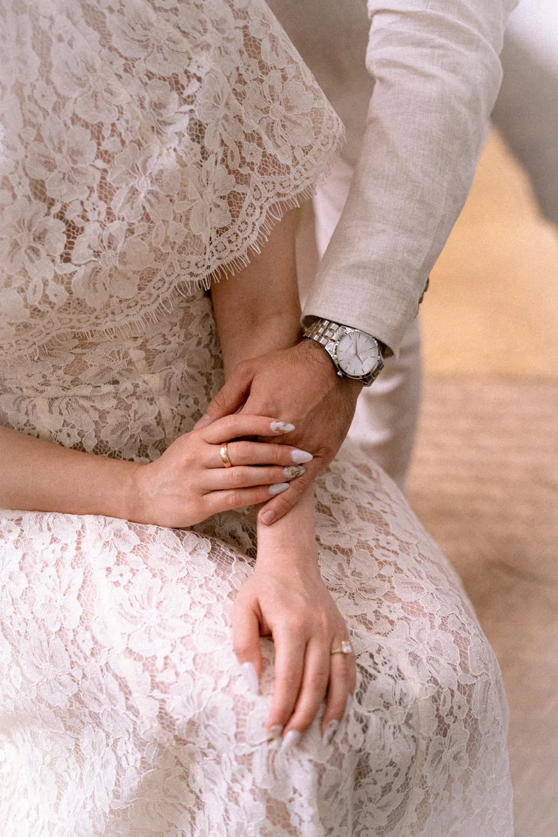 Las manos de los novios en reposo sobre el encaje del vestido, con los anillos y el reloj