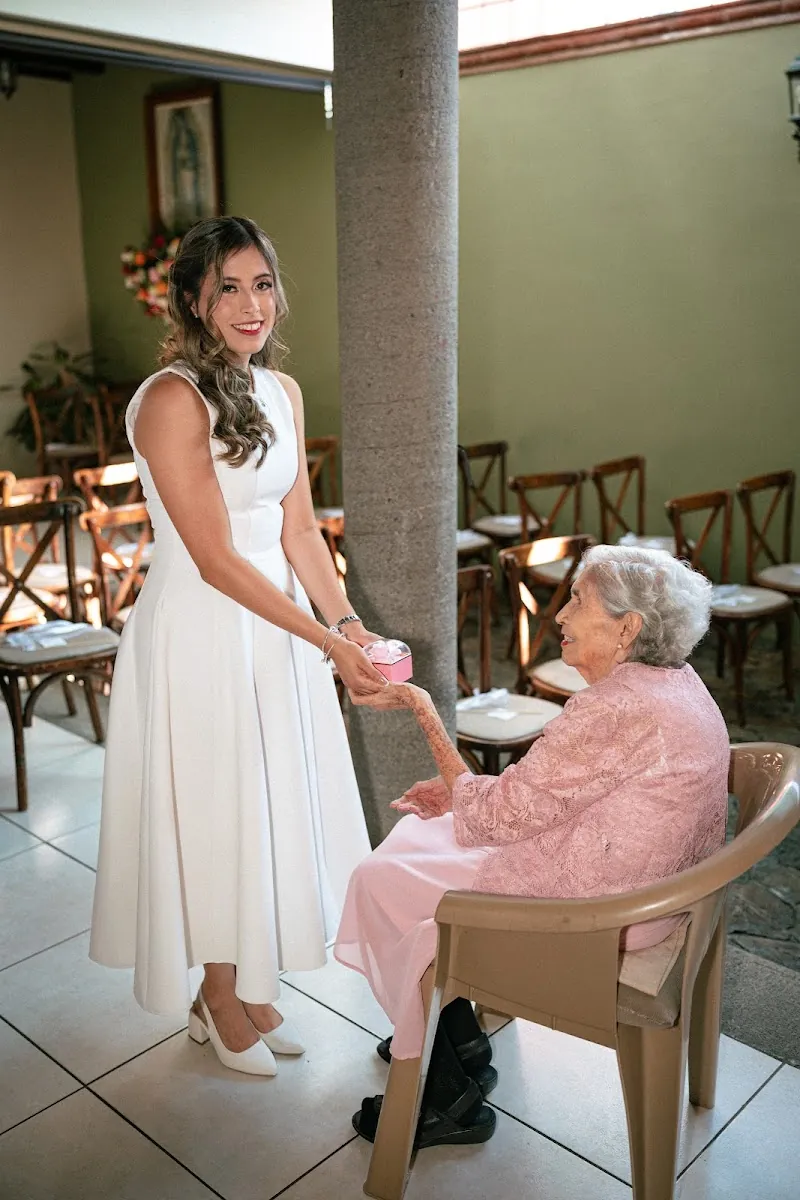 La novia, de pie y sonriendo, toma la mano de su abuela sentada